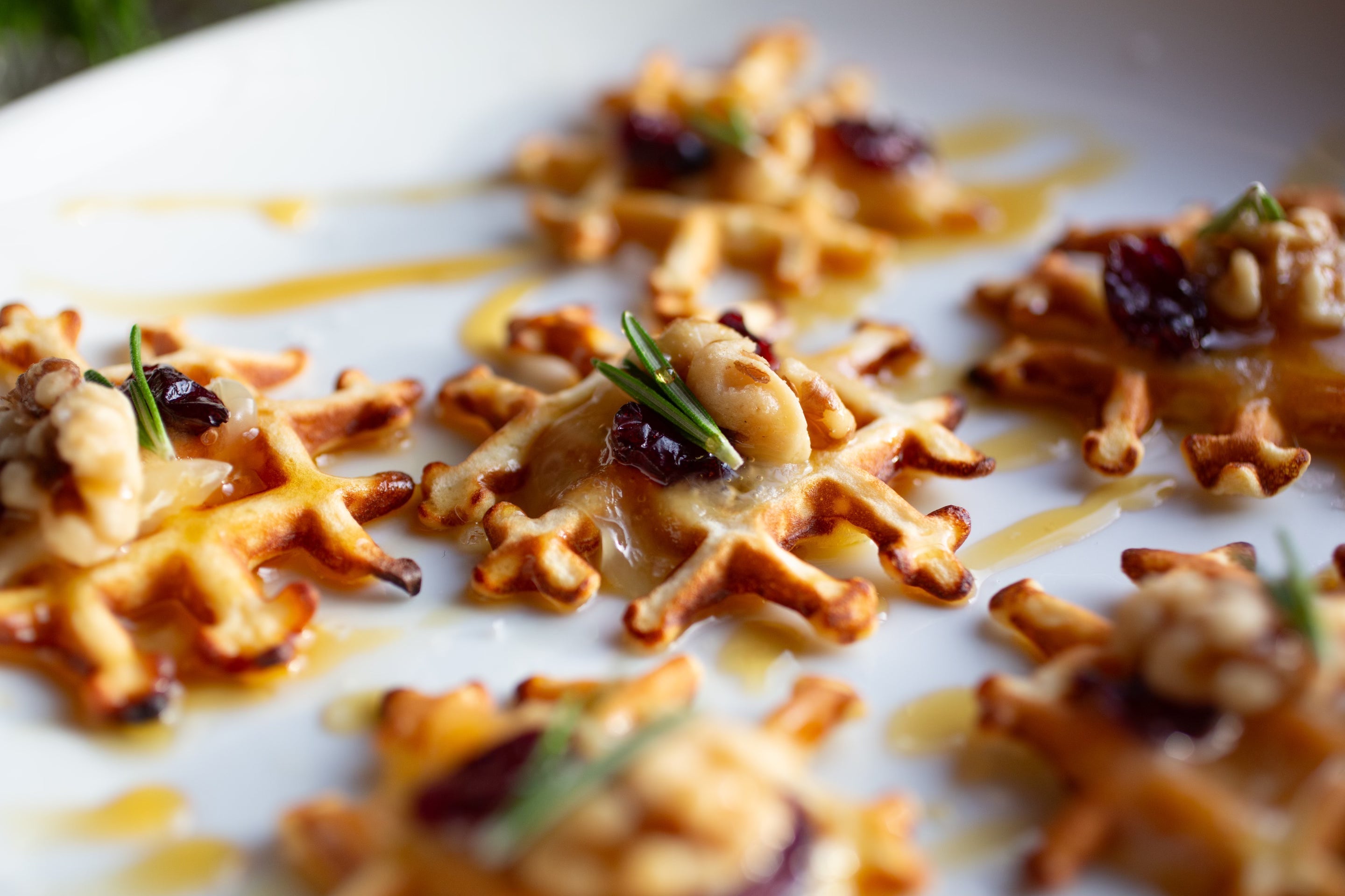 Heavenly Waffles Holiday Brie Bites topped with melted brie, dried cranberries, walnuts, rosemary, and maple syrup on a serving platter.